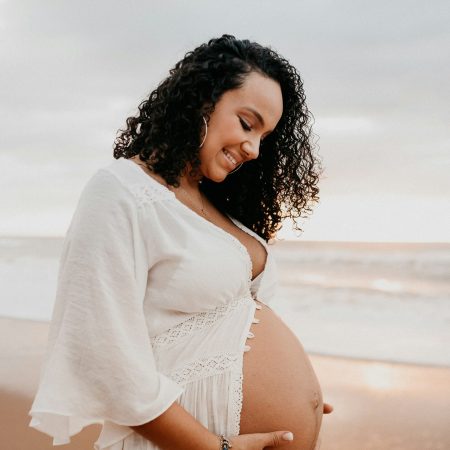 A pregnant woman in a flowing white dress stands on a sandy beach.