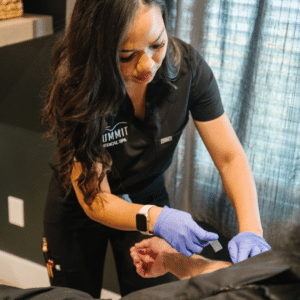A spa professional in a black uniform applies a treatment to a client's wrist, wearing blue gloves in a serene setting.