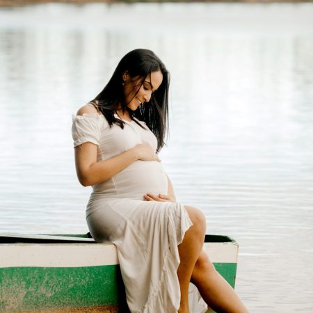 A pregnant woman in a flowing white dress sits on the edge of a boat, gazing thoughtfully at the water, surrounded by natural beauty.