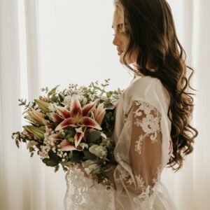 A woman in a lace-trimmed robe holds a bouquet of lilies and greenery, standing by a window with sunlight filtering through.