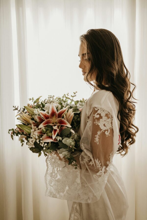 A woman in a lace-trimmed robe holds a bouquet of lilies and greenery, standing by a window with sunlight filtering through.